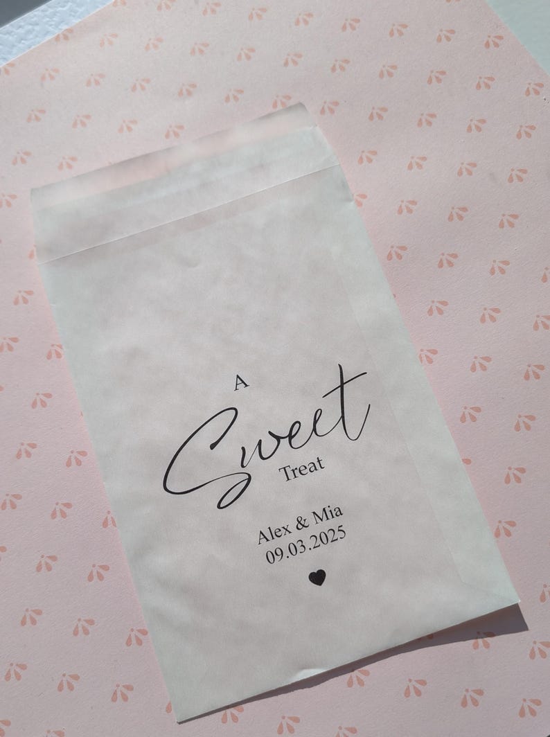 May include: A white paper bag with the text "A Sweet Treat" in black cursive font. Below the text is "Alex & Mia" and the date "09.03.2025" in black font. A small black heart is below the date.