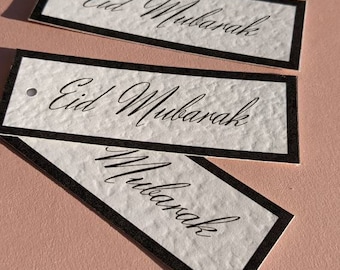 May include: A stack of white gift tags with black borders and a hole punched in the top. The tags have the text "Eid Mubarak" written in black calligraphy.