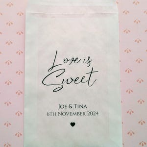 May include: White paper favour bag with the text "Love is Sweet" in black cursive font. Below the text is "Joe & Tina" and "6th November 2024" in black font. A small black heart is below the date.