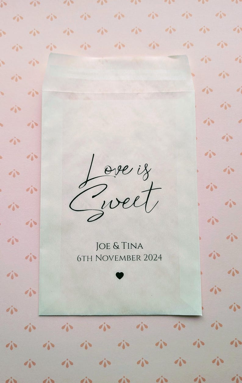 May include: White paper favour bag with the text "Love is Sweet" in black cursive font. Below the text is "Joe & Tina" and "6th November 2024" in black font. A small black heart is below the date.
