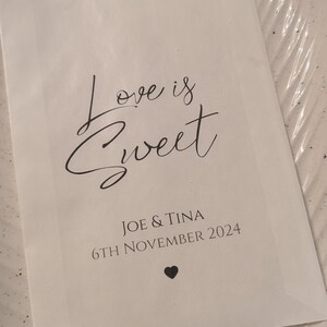 May include: White paper bag with the text "Love is Sweet" in black cursive font. Below the text is "Joe & Tina" and "6th November 2024" in black font. A small black heart is at the bottom of the bag.