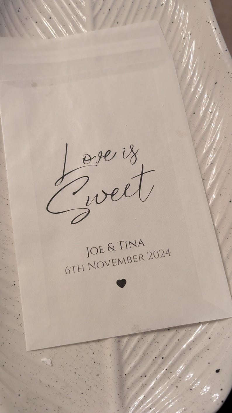 May include: White paper bag with the text "Love is Sweet" in black cursive font. Below the text is "Joe & Tina" and "6th November 2024" in black font. A small black heart is at the bottom of the bag.