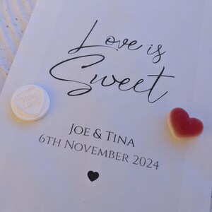 May include: White paper bag with the text "Love is Sweet" in black cursive font. Below the text is "Joe & Tina" and "6th November 2024". There are three heart-shaped sweets on the bag: one white, one red, and one yellow.