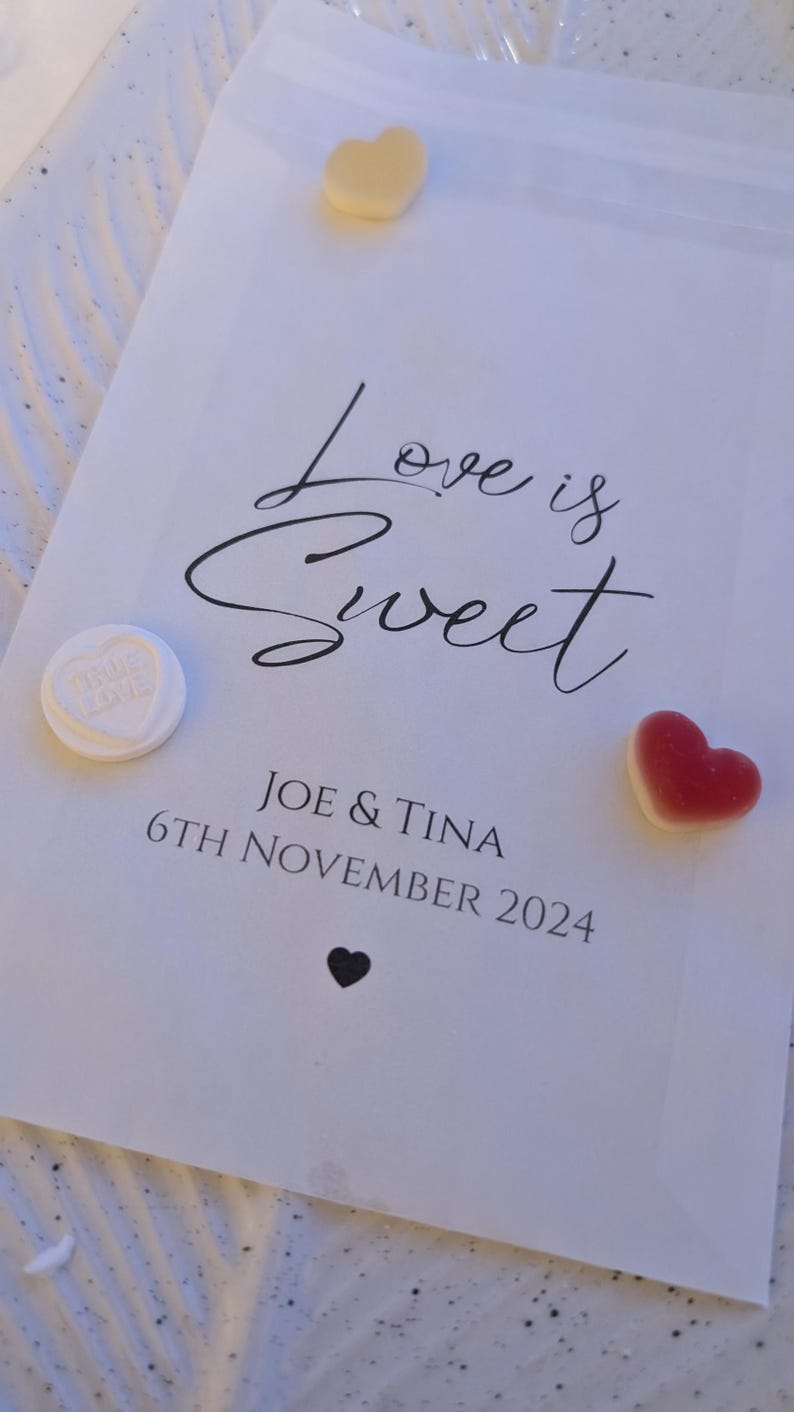 May include: White paper bag with the text "Love is Sweet" in black cursive font. Below the text is "Joe & Tina" and "6th November 2024". There are three heart-shaped sweets on the bag: one white, one red, and one yellow.