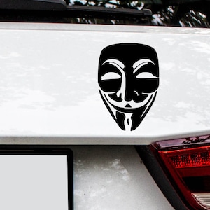 Puede incluir: Una pegatina en blanco y negro de la máscara de Anonymous en la parte trasera de un coche blanco.