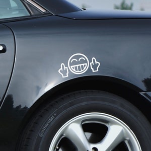 Peut inclure: Une voiture noire avec un autocollant de visage souriant blanc avec deux doigts d'honneur sur l'aile arrière.
