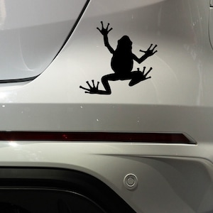Op de afbeelding: Een zwarte kikkersticker is bevestigd aan de achterkant van een zilverkleurig voertuig. De kikker wordt afgebeeld als silhouet, met uitgestrekte ledematen en zwemvliezen, wat beweging suggereert. Een decoratief accessoire.