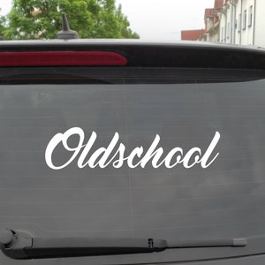 Puede incluir: Una calcomanía de vinilo blanco con la palabra "Oldschool" escrita en letra cursiva en la ventana trasera de un coche negro.