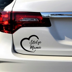 以下が含まれることがあります： 黒いビニール製の車のステッカー。ハートの形をしており、筆記体で「Stolze Mama」と書かれています。ステッカーは、車の白い後部、テールライトの近くに貼られています。