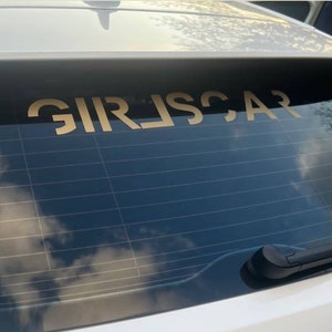 May include: A white car with a black rear window. The rear window has a gold decal that reads "GIRL'S CAR".