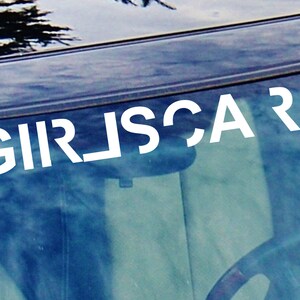 May include: A white vinyl decal with the text "GIRLSCAR" on a car windshield.