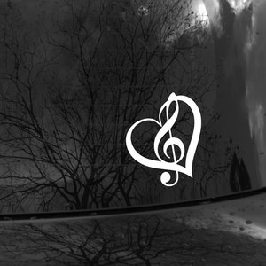 Puede incluir: Calcomanía de vinilo blanco de una clave de sol dentro de una forma de corazón. La calcomanía está en una ventana de automóvil negra.