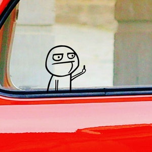 Könnte beinhalten: Ein schwarz-weißer Meme-Aufkleber eines Strichmännchens, das den Mittelfinger zeigt, durch ein Autofenster sichtbar. Der Aufkleber befindet sich auf der Scheibe eines roten Autos. Das Strichmännchen hat einen selbstgefälligen Gesichtsausdruck.