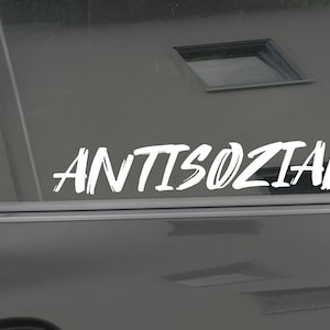 以下が含まれることがあります： 太字でスタイリッシュなフォントで「ANTISOZIAL」と書かれた白いビニールステッカー。ステッカーは車のリアウィンドウに貼られています。