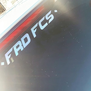 May include: A close-up of a white sticker with the text "- FRD FCS -" on a black surface.
