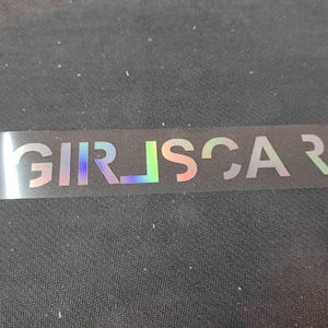 May include: A clear sticker with the iridescent text "GIRLSCAR" printed on it.