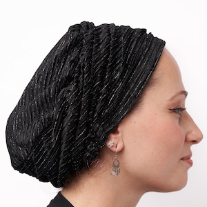 May include: A black head covering with a textured, pleated design. The fabric has subtle silver threads woven throughout, adding a touch of shimmer. The head covering is worn by a person, and the image is set against a white background.