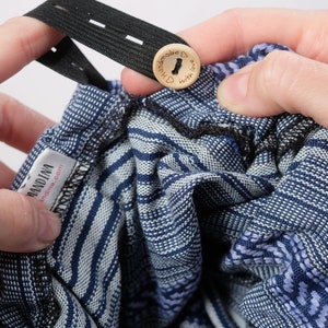 May include: A blue and white patterned baby wrap with a black elastic strap and a wooden button that says "Handmade with love". The wrap has a label that says "Mandina" and "mandina.com".