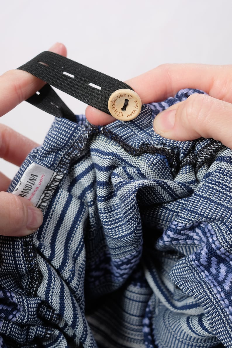 May include: A blue and white patterned baby wrap with a black elastic strap and a wooden button that says "Handmade with love". The wrap has a label that says "Mandina" and "mandina.com".