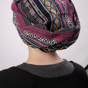 May include: A black, pink, blue, and yellow patterned turban style head wrap. The head wrap is made of a soft, woven fabric.