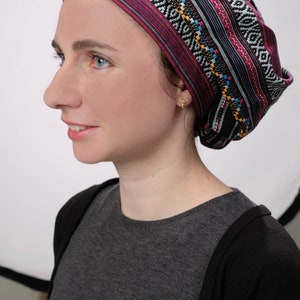 May include: A woman wearing a black and pink patterned head covering with a geometric design. The head covering is tied in a knot at the back of her head. She is wearing a gray shirt and a black cardigan.