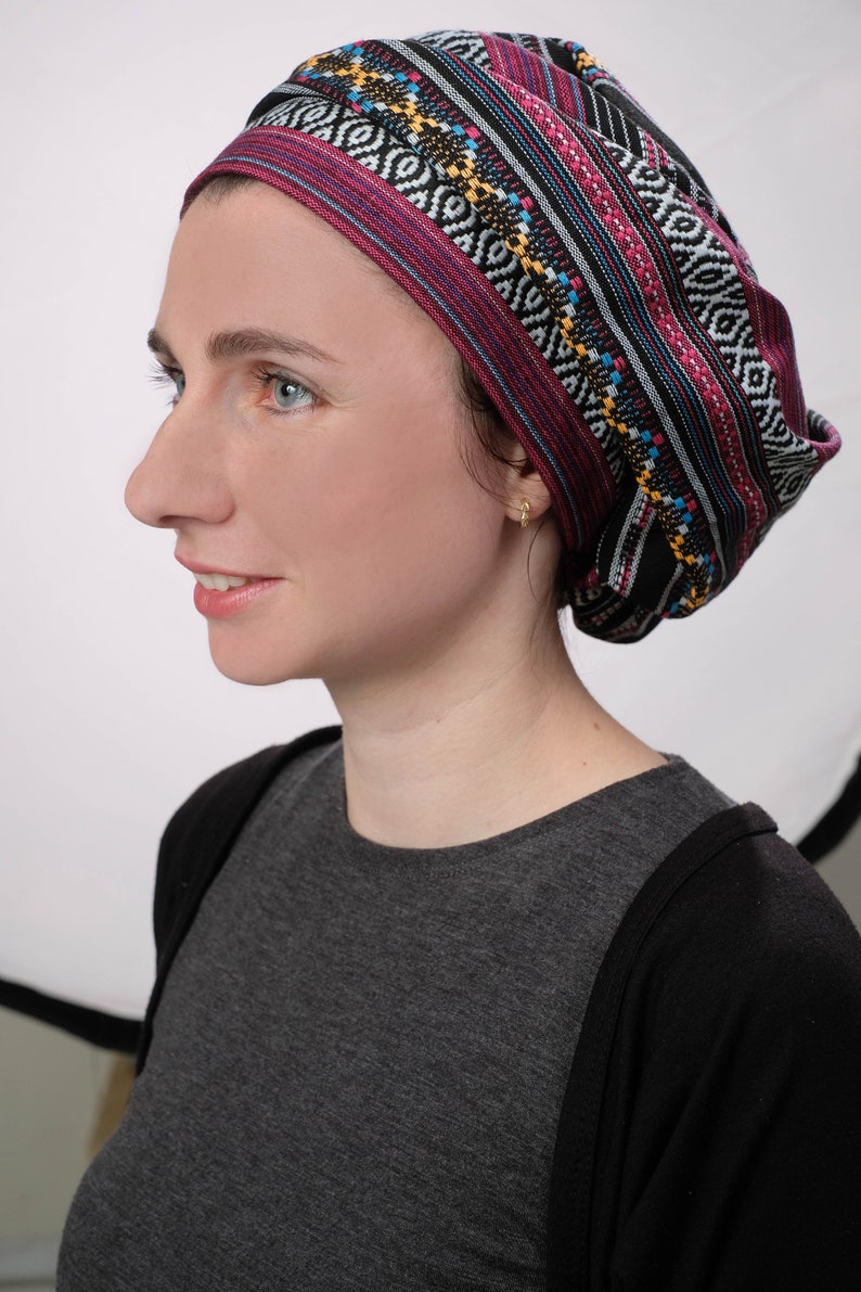 May include: A woman wearing a black and pink patterned head covering with a geometric design. The head covering is tied in a knot at the back of her head. She is wearing a gray shirt and a black cardigan.