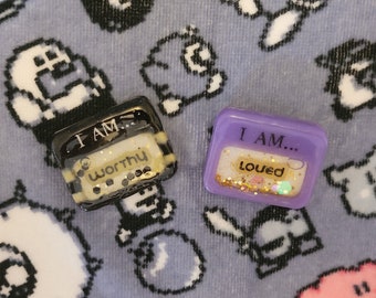 May include: Two rectangular resin pins with rounded corners. The black pin features the text I AM... worthy with embedded glitter. The purple pin features the text I AM... loved with embedded gold and iridescent glitter accents inside the resin.