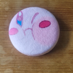 Puede incluir: Un juguete de peluche rosa redondo que representa un personaje de dibujos animados con mejillas rosas, boca rosa y un ojo azul y blanco. El juguete está sobre una superficie de madera.
