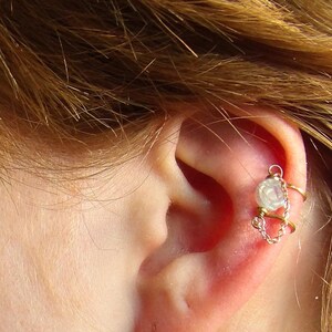 May include: A delicate ear cuff featuring a small, round, white gemstone and a gold-colored metal design. The cuff wraps around the ear, with a chain detail connecting the top and bottom sections. The image shows the ear and part of the face.