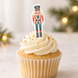 Op de afbeelding: Een vanillecupcake met witte glazuur, versierd met kleine gouden en witte strooisels, bekroond met een platte, geïllustreerde papieren notenkraker-topper in een rood, blauw en groen uniform met gouden accenten in het midden.