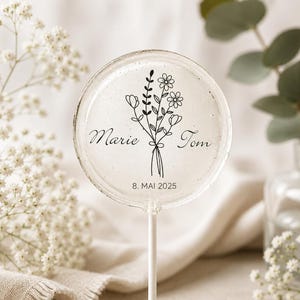 May include: A round, clear lollipop featuring a black line-art illustration of a wildflower bouquet. The text "Marie" and "Tom" is printed on either side of the flowers, with the date "8. MAI 2025" printed in black text at the bottom.