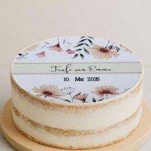 Puede incluir: Una tarta redonda con un diseño floral en la parte superior, con flores rosas y crema y hojas verdes. La tarta tiene un glaseado de color claro y se muestra en un soporte de madera para tartas. El texto en la tarta dice "Taufe von Emma 10. Mai 2026."