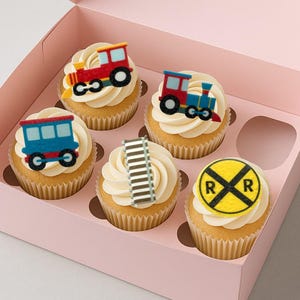 Puede incluir: Seis cupcakes en una caja rosa, cada uno cubierto con crema de vainilla. Los cupcakes están decorados con decoraciones comestibles con temas de trenes y ferrocarriles, incluyendo vagones y una señal de cruce ferroviario.