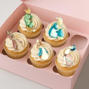 Puede incluir: Seis cupcakes con glaseado blanco y decoraciones comestibles de personajes, incluyendo un zorro, conejos y un pato, están dispuestos en una caja rosa. Los cupcakes tienen una base marrón claro y están en moldes de papel. La caja está abierta.