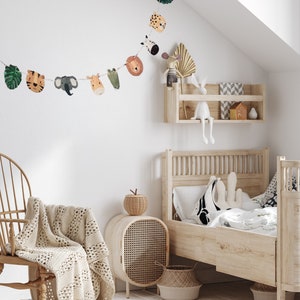 May include: A white wooden bed with a white and green floral patterned bedding. A white crocheted throw blanket is draped over a wooden chair. A wooden side table with a woven rattan door and a woven basket sits next to the chair. A woven basket sits at the foot of the bed. A garland of animal faces hangs on the wall above the bed.