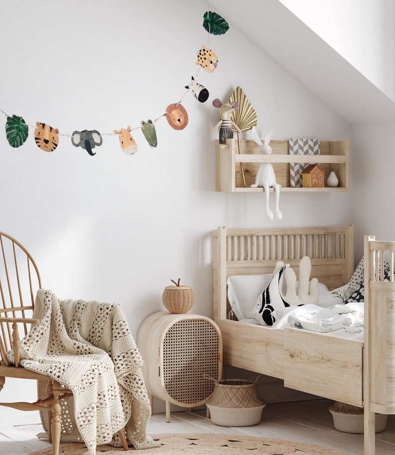 May include: A white wooden bed with a white and green floral patterned bedding. A white crocheted throw blanket is draped over a wooden chair. A wooden side table with a woven rattan door and a woven basket sits next to the chair. A woven basket sits at the foot of the bed. A garland of animal faces hangs on the wall above the bed.