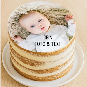 Può includere: Una torta rotonda con una foto di un bambino e la scritta "DEIN FOTO & TEXT". La torta ha diversi strati di pan di spagna e crema, presentata su un piatto bianco. Poggia su una superficie di legno marrone chiaro.