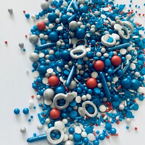 May include: A mix of blue, red, silver, and white sprinkles. The sprinkles are in the shape of balls, stars, and rings.