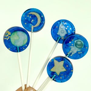 Puede incluir: Cinco piruletas azules con diseños espaciales, incluyendo una luna creciente, la Tierra, un cohete, un astronauta y una estrella. Cada piruleta tiene un palito blanco y detalles de estrellas doradas. Las piruletas se sostienen en una mano sobre un fondo blanco.