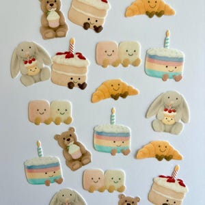 Peut inclure: Une collection de décorations d'anniversaire fantaisistes en feutre. L'image présente des ours en peluche souriants, des lapins, des gâteaux avec des bougies, des croissants et des paires de formes rectangulaires souriantes. La palette de couleurs comprend des pastels et des tons chauds.