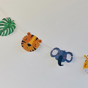 May include: A green leaf, a tiger face, a gray elephant face, and a yellow giraffe face are strung together on a white string. This is a jungle-themed party banner.