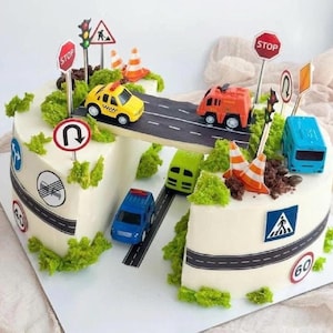 May include: A decorated cake with a road theme. The cake features a road design with toy cars, traffic cones, and road signs. The cake is decorated with green frosting to resemble bushes and grass.