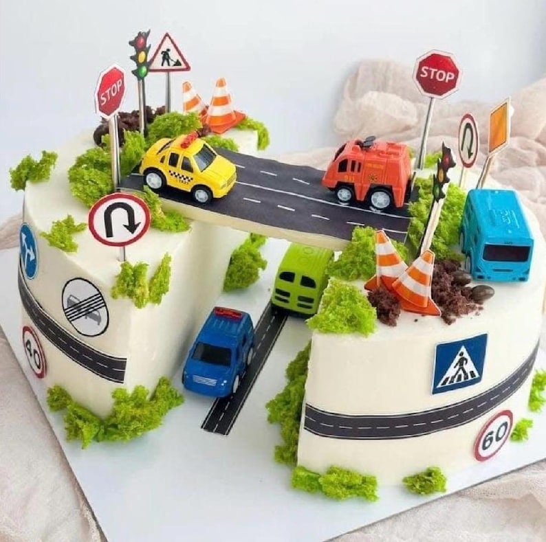 May include: A decorated cake with a road theme. The cake features a road design with toy cars, traffic cones, and road signs. The cake is decorated with green frosting to resemble bushes and grass.
