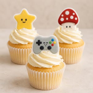 May include: Three cupcakes with white frosting, each topped with a flat edible decoration. One features a yellow smiling star, one a red and white spotted mushroom, and the centre one a grey video game controller with colourful buttons.