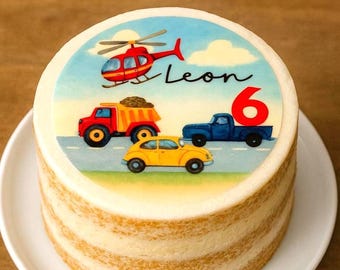 May include: A round birthday cake with a colorful design featuring a red helicopter, a red dump truck, a yellow car, and a blue truck. The cake has the name "Leon" and the number "6" on top. The cake is on a white plate.