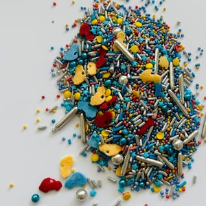May include: A colorful assortment of sprinkles, including silver rods, blue, red, and yellow shapes, and small round sprinkles. The sprinkles are scattered on a white surface, creating a vibrant and festive display, perfect for decorating baked goods.