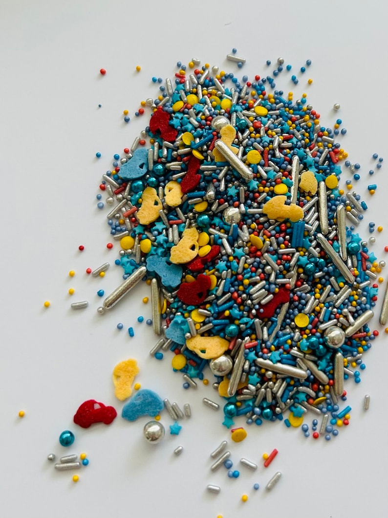 May include: A colorful assortment of sprinkles, including silver rods, blue, red, and yellow shapes, and small round sprinkles. The sprinkles are scattered on a white surface, creating a vibrant and festive display, perfect for decorating baked goods.