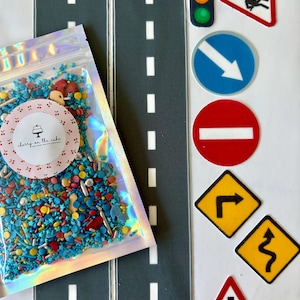 May include: An assortment of cake decorating supplies, including two gray road mats with white dashed lines, a bag of colorful sprinkles, and various road sign cutouts. The bag has a label that says "cherry on the cake".