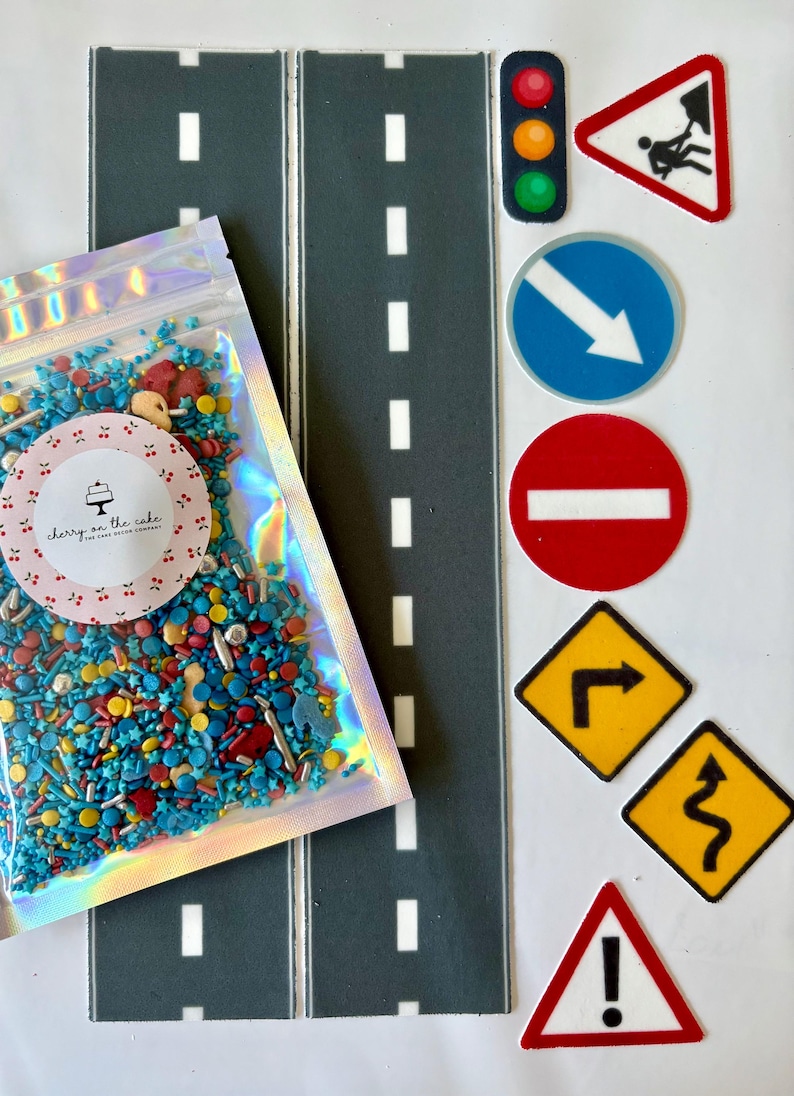 May include: An assortment of cake decorating supplies, including two gray road mats with white dashed lines, a bag of colorful sprinkles, and various road sign cutouts. The bag has a label that says "cherry on the cake".