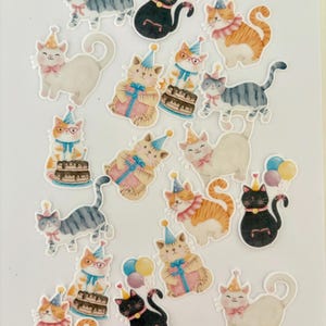 Puede incluir: Una colección de pegatinas de gatos coloridos en varias poses, con sombreros de fiesta y lazos. Algunos gatos sostienen regalos, globos o están cerca de pasteles. Los gatos son de color gris, naranja, negro y blanco, con acentos rosas y azules.